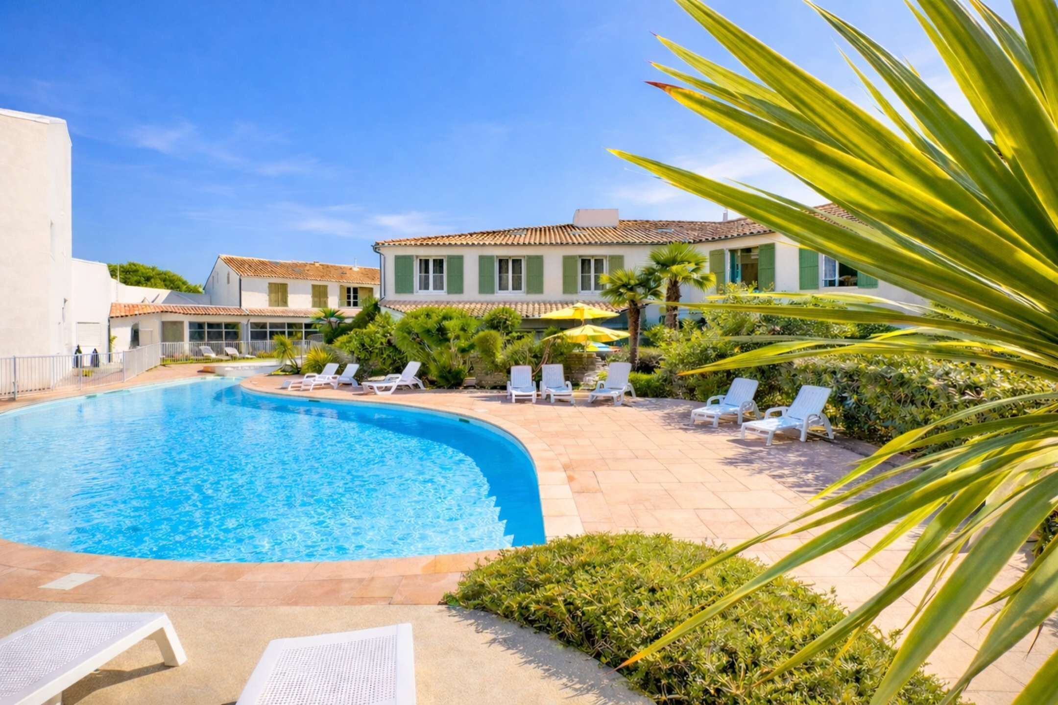 Une piscine avec des chaises longues, un bâtiment aux volets verts et un ciel bleu azur.