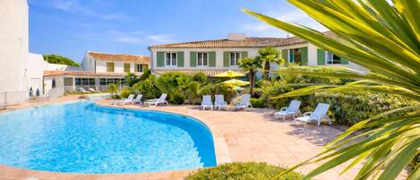 A swimming pool with lounge chairs, a building with green shutters, and a clear blue sky.