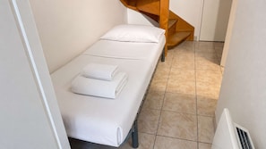 A small, single-bed room with a white mattress, a neatly folded white towel, and a wooden staircase in the background. - Apartment with communal pool and parking (LA COUARDE SUR MER)