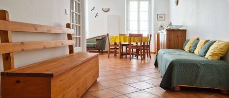 A room with a wooden bench, a dining table with chairs, a bed with pillows, and a window.