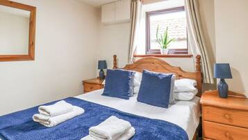 A neatly made bed with pillows, a blue blanket, and two white towels. There is a wooden nightstand with a lamp and a potted plant on the windowsill.