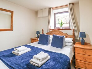A neatly made bed with pillows, a blue blanket, and two white towels. There is a wooden nightstand with a lamp and a potted plant on the windowsill. - Owl (Pickering)