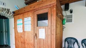 Eine Holzsauna mit Informationsplakaten, einer Tür, einem Fenster, einer Lüftungsanlage und einem Plastikstuhl.
