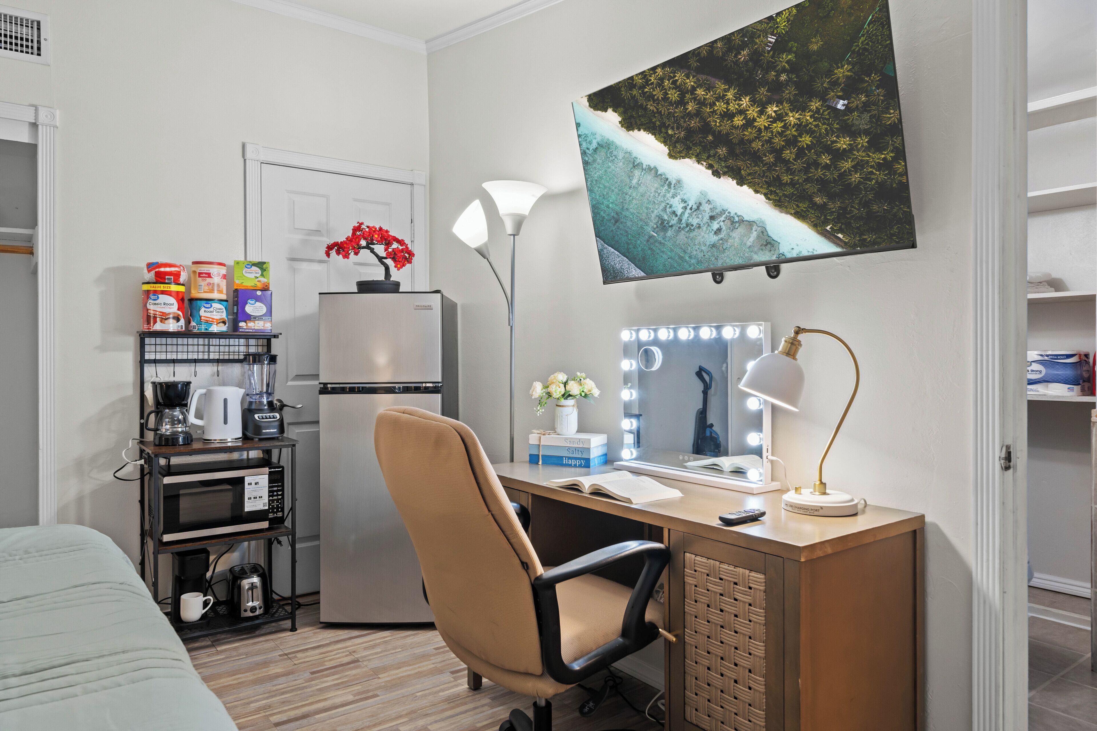 A hotel room with a kitchenette, a bed, a desk with a computer and a lamp, and a large window with a view of a beach.