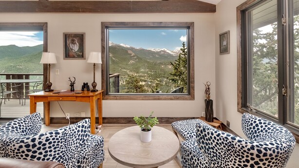 Ein Zimmer mit großem Fenster und Blick auf die Berge, einem runden Tisch mit Topfpflanze, Leopardenmuster-Stühlen und einem Holztisch mit Lampen.