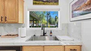 Una cocina con un lavaplatos de acero inoxidable, armarios de madera, un escurreplatos y una ventana con vistas a árboles y un edificio.