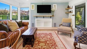 Una sala de estar con un sofá de cuero, una mesa de centro de madera, un tapete estampado, un televisor montado en la pared y una silla con cojín estampado.