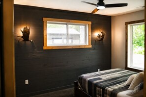 A bedroom with a dark wooden wall, a window with a wooden frame, a ceiling fan, and a bed with a striped bedspread. - Modern Rustic Cabin - ATV & Snowmobile Trail Access - Fire Pit (Hayward)