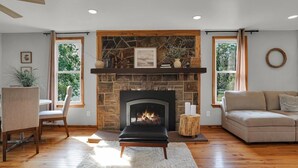 A living room with a stone fireplace, wooden flooring, a sofa, a dining table with chairs, and large windows. - Riverfront Manor - Beautiful view and setting! (Dauphin)