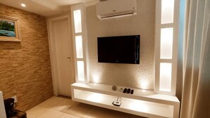 A modern living room with a flat-screen TV mounted on a white wall, a built-in shelf below, and a window with frosted glass panels. - Pousada Requinte da Serra  (Miguel Pereira)