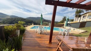 A wooden deck with a pool, chairs, and a pergola overlooking a mountainous landscape. - Pousada Requinte da Serra  (Miguel Pereira)