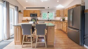 Een moderne keuken met houten kastjes, een centraal eiland met barkrukken, een roestvrijstalen koelkast, en een raam met uitzicht op groen.
