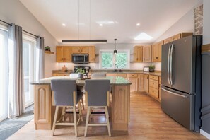 A modern kitchen with wooden cabinets, a central island with bar stools, a stainless steel refrigerator, and a window with a view of greenery. - NH Escape, 4 Bedroom, Pool, Hot Tub, Quiet Location (Goffstown)
