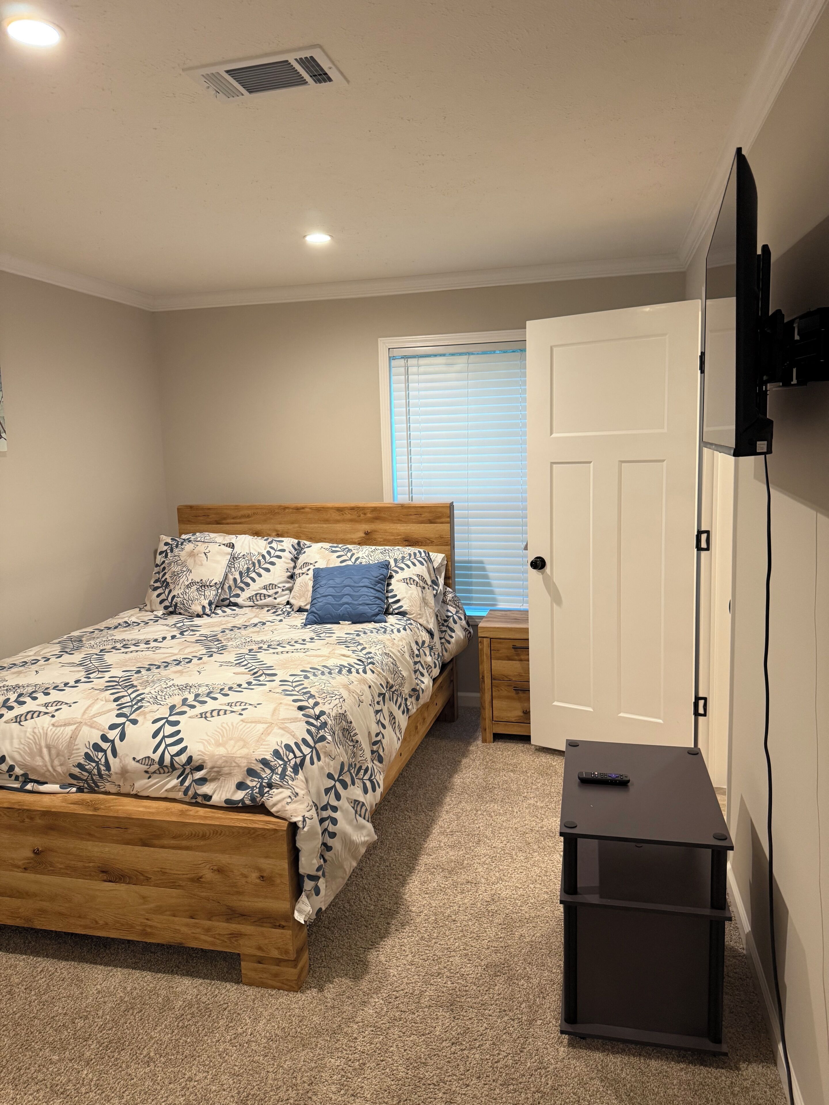 A bedroom with a wooden bed, a bedside table, a TV mounted on the wall, and a window with blinds.
