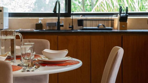 A dining area with a table set for two, a window with a view of greenery, and a kitchen area with cabinets and a sink. - Depto. Eek’ · 1 Hab · 1 Baño · Terraza Privada (Merida)