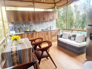 Una acogedora cocina al aire libre con armarios de madera y un salpicadero de mármol, una mesa de comedor con mantel estampado, y un sofá de mimbre con cojines.