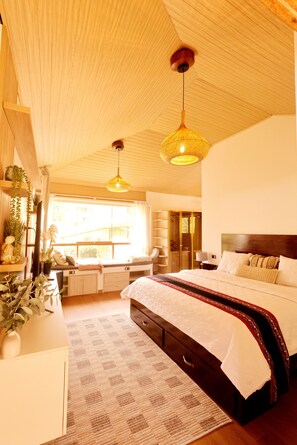A bedroom with a large bed, a window with a view, a wooden ceiling, and a hanging light fixture.