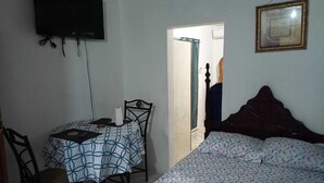 Un dormitorio con cama, una mesa con mantel estampado, una silla, televisor montado en la pared y un cuadro enmarcado.