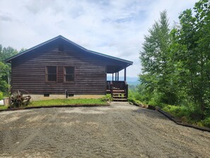 A rustic wooden cabin with a porch, surrounded by lush greenery and a gravel driveway.