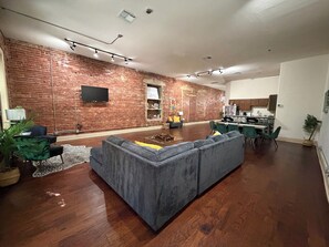 A modern living room with a brick wall, a grey sectional sofa, and a dining area with green chairs. - The Safari Unit 2 Downtown Loft Sleeps 4 (Oklahoma City)