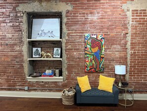 A room with a brick wall, a sofa with yellow cushions, a lamp, and a shelf with decorative items. - The Safari Unit 2 Downtown Loft Sleeps 4 (Oklahoma City)