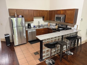 A kitchen with wooden cabinets, stainless steel appliances, a granite countertop, and a bar area with stools. - The Safari Unit 2 Downtown Loft Sleeps 4 (Oklahoma City)
