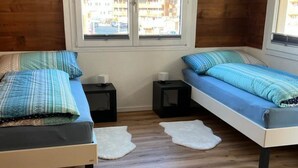 Ein Zimmer mit zwei Betten, einem kleinen Tisch und einem Stuhl. Zwei Fenster bieten Blick auf umliegende Gebäude.