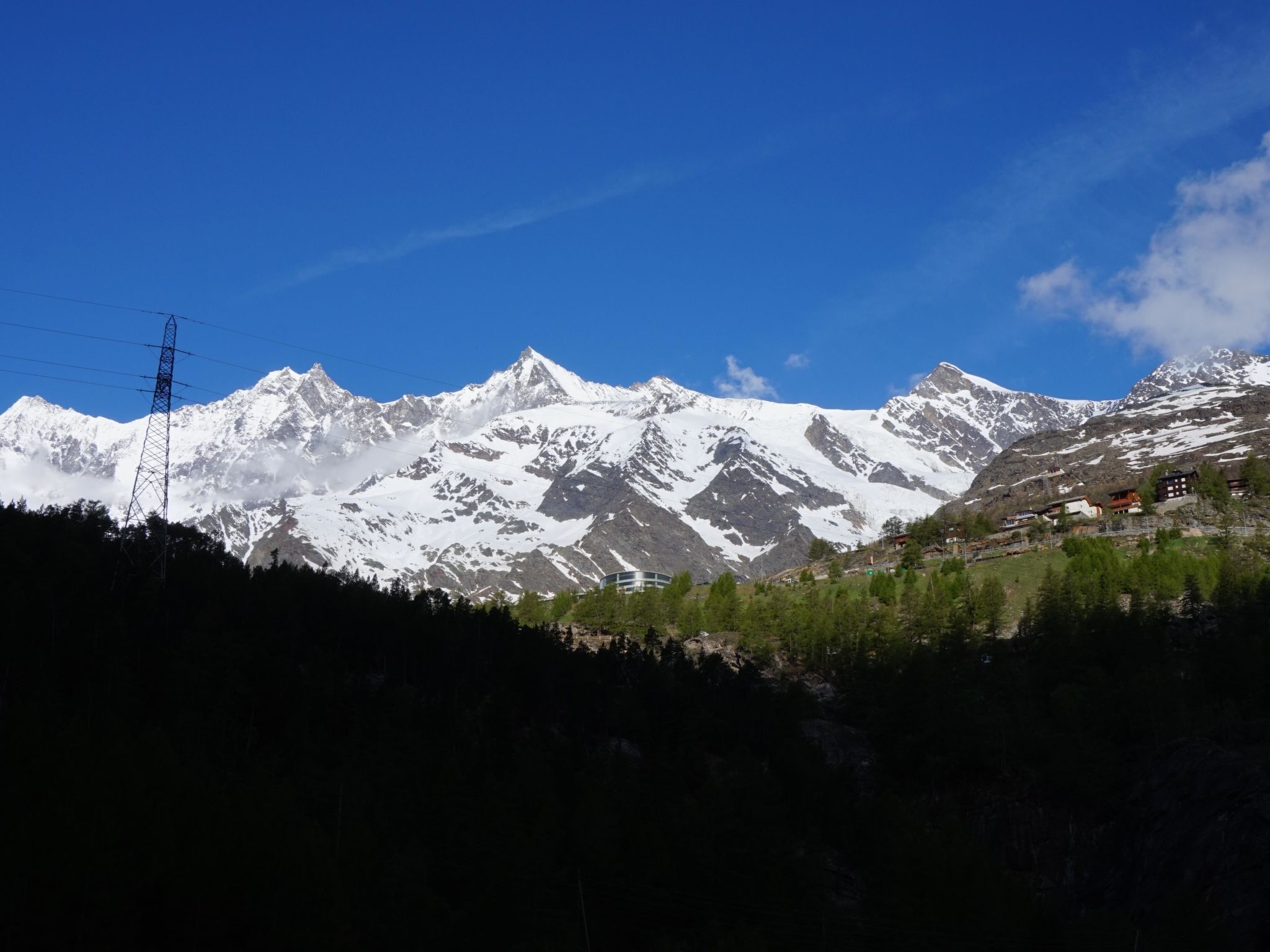 Paisagem montanhosa com picos nevados, uma linha de energia e um pequeno povoado com edifícios.