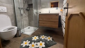 Een moderne badkamer met een glazen douchecabine, een wit toilet, een houten wastafelmeubel en een grijze vloerkleed met witte en gele bloemen.