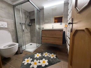 Ein modernes Badezimmer mit einer Duschkabine aus Glas, einer weißen Toilette, einem Holzwaschtisch und einem grauen Teppich mit weißen und gelben Blumen.