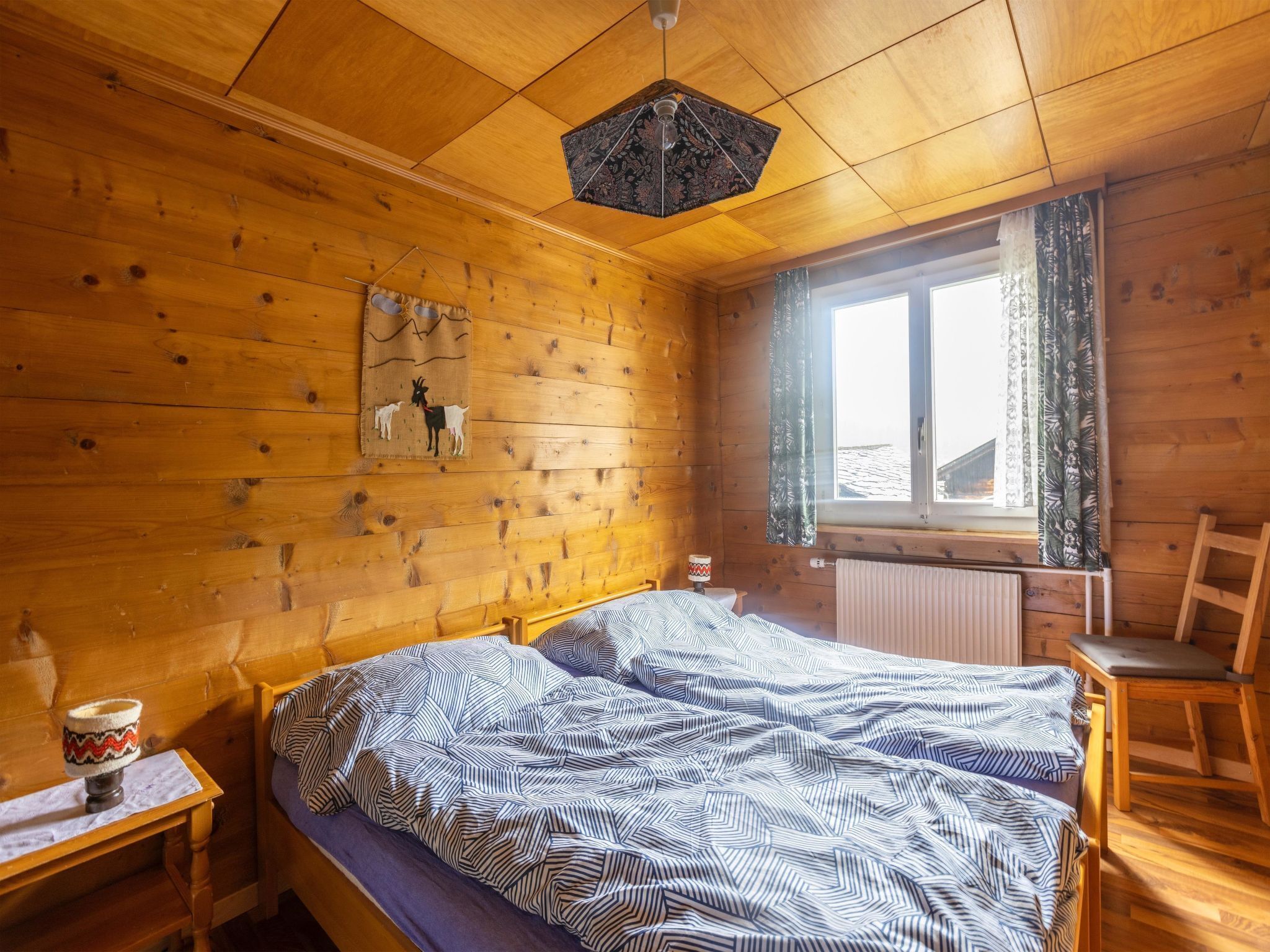 A wooden cabin bedroom with a bed, a nightstand, a chair, and a window with curtains.