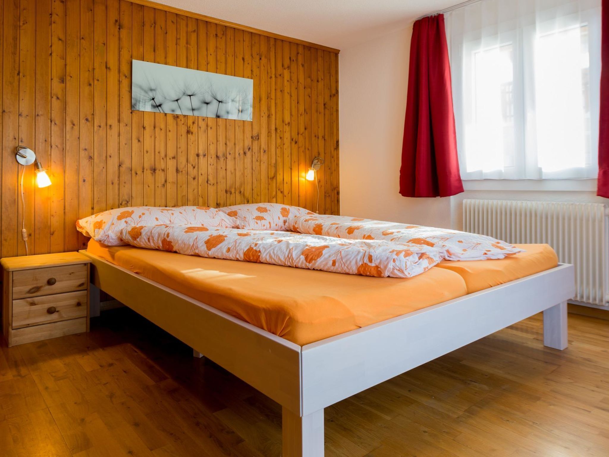 Une chambre avec un mur en bois, un lit recouvert d’une couverture orange, une table de chevet, deux lampes fixées au mur et une fenêtre avec des rideaux rouges.