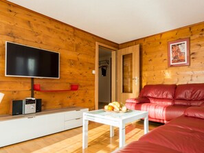 A cozy living room with a wooden wall, a flat-screen TV, a red leather sofa, and a small table with fruit. - Chalet Bambi Appartement/Fewo 6-Bettwohnung Nr. 1 by Interhome (Saas-Fee)