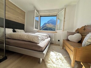 A bedroom with a bed, a wooden dresser, a fluffy rug, and a window with a mountain view. - Bettina Wellness Appartement/Fewo, Dusche oder Bad Bettina 3 by Interhome (Saas-Fee)