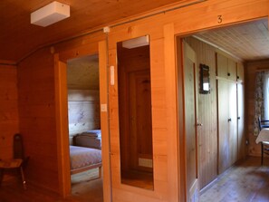 Interior de madeira com uma cama, uma cadeira e uma mesa. Há portas que dão para outros cômodos.