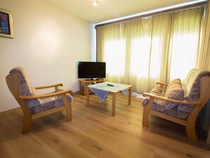 Een woonkamer met twee houten stoelen, een bijzettafeltje, een televisie en een raam met gordijnen.