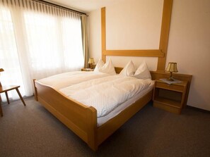 Een hotelkamer met een tweepersoonsbed, twee nachtkastjes met lampen, een stoel en een groot raam met gordijnen.
