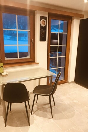 A dining area with a table and two chairs, a window with a view of a snowy landscape, and a door with a glass panel. - Art Oase Appartement/Fewo 2-Bettwohnung by Interhome (Saas-Grund)