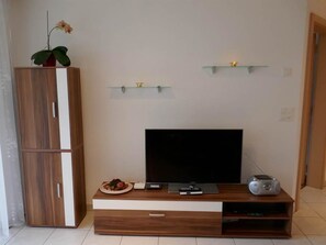 A living room with a flat-screen TV, wooden entertainment unit, a small table with a plate of food, a radio, and a potted plant. - Haus Artemis Appartement/Fewo 5-Bettwohnung Nr. 51 by Interhome (Saas-Grund)
