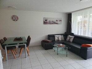 A modern living room with a dining table, a glass coffee table, a black sofa, and a wall clock. - Haus Artemis Appartement/Fewo 5-Bettwohnung Nr. 51 by Interhome (Saas-Grund)