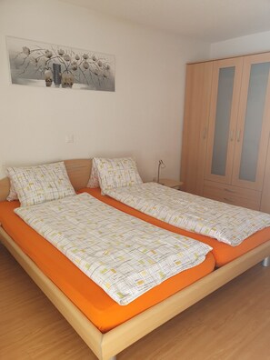Cama doble con colchón naranja y ropa de cama blanca con diseño geométrico. A la derecha, un armario de madera con puertas de vidrio. En la pared, encima de la cama, un cuadro con un árbol y flores.