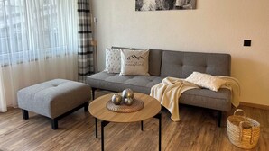 Una sala de estar con un sofá gris, un reposapiés gris, una mesa de centro redonda de madera con adornos, y un cuadro en blanco y negro en la pared.