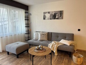 Una sala de estar con un sofá gris, un reposapiés gris, una mesa de centro redonda de madera con adornos, y un cuadro en blanco y negro en la pared.