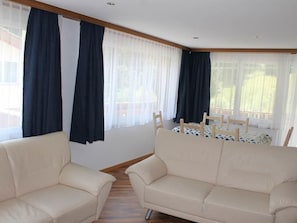 Ein Zimmer mit zwei hellbeigen Sofas, die auf einen Esstisch mit Stühlen zeigen, großen Fenstern mit dunklen Vorhängen und Holzboden.