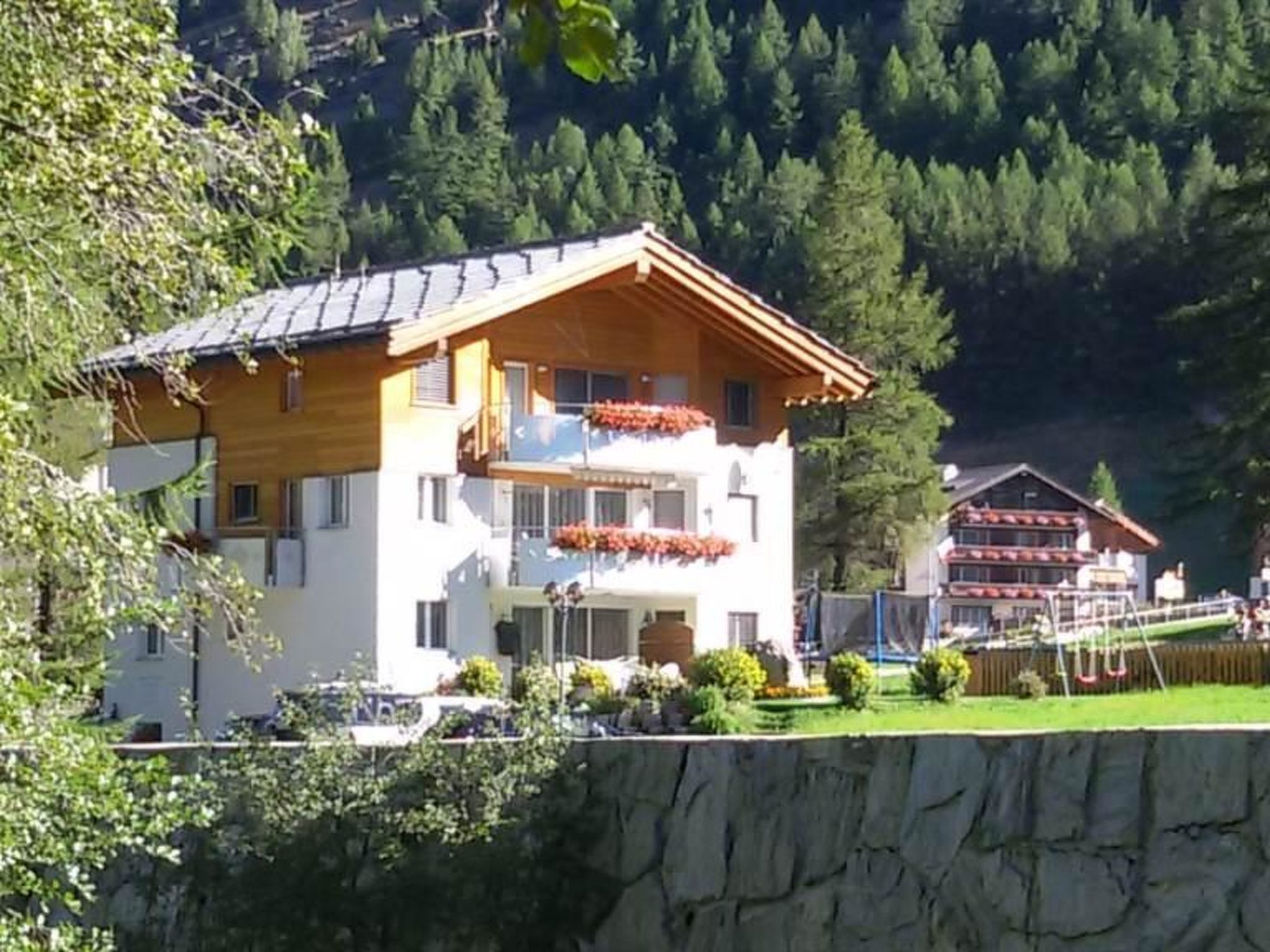 Une maison en bois à deux étages, dotée d’un balcon, entourée de verdure et située au pied d’une colline rocheuse.