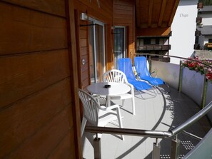 Een balkon met een tafel, stoelen en een glazen hekwerk, en een gebouw met een houten gevel.