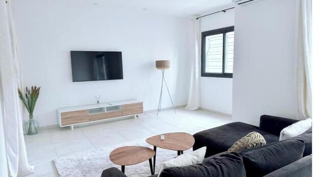 A modern living room with a large flat-screen TV mounted on the wall, a floor lamp, a sofa, and a coffee table. - Spacious Two-Bedroom Apartment with a Private Balcony and Terrace (Dakar)