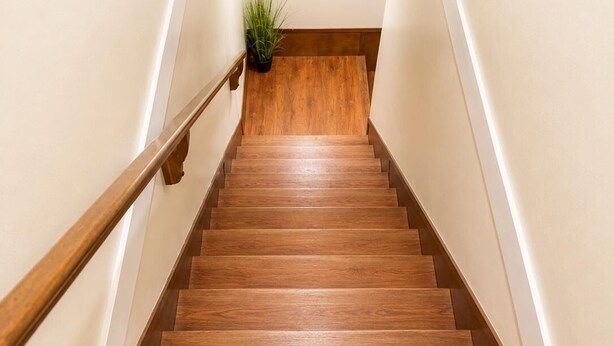 Eine Holztreppe mit Handlauf, weißen Wänden und einer kleinen Pflanze oben.