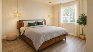 Un dormitorio con una cama, dos lámparas colgantes, una ventana con cortinas, una planta en maceta y un otoman redondo.