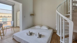 Hotelkamer met een bed, witte kussens, een witte stoel, een bureau, een lamp en een wenteltrap.